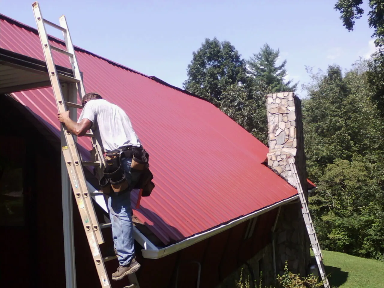 Roofer on a ladder beside a red metal roof and stone chimney — Soffit & Fascia Repair by Roofing Brothers in Sciota